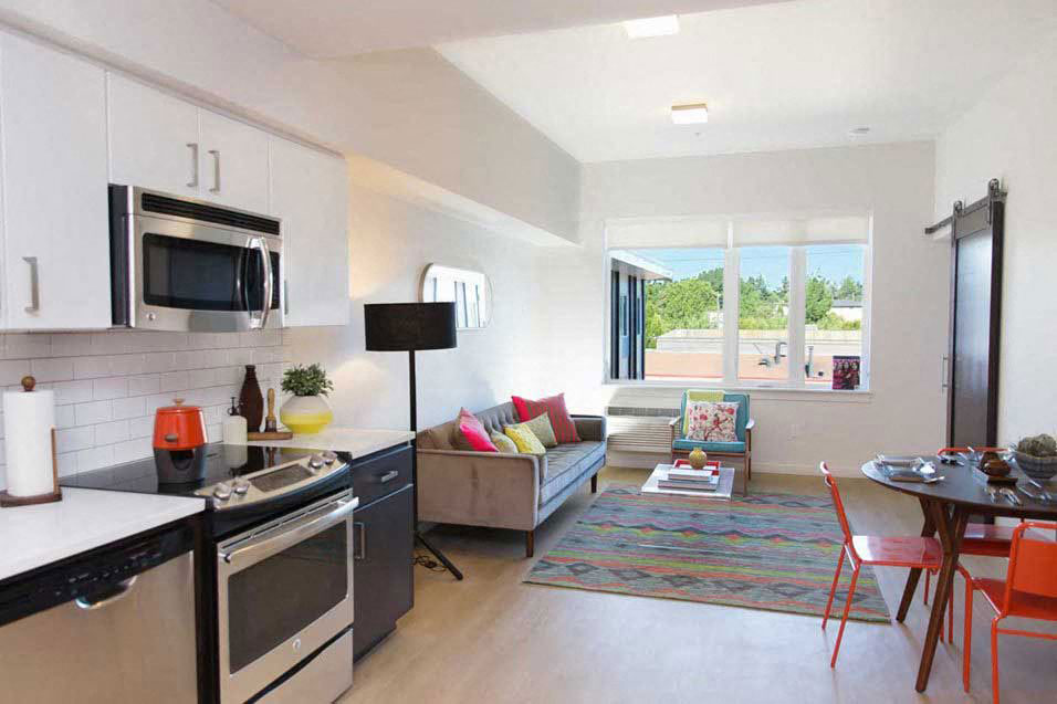 A modern kitchen with a black stove top oven and microwave, a dining table with chairs, and a couch with colorful pillows.