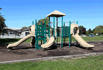 Outdoor playscape with 3 slides and green climbing area in center.  On woodchips surrounded by green grass.