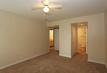Empty bedroom with a white ceiling fan with light in center.  Brown carpet, walk in closet off bedroom.