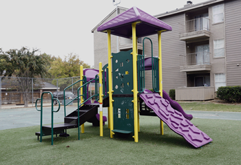 Green playscape with yellow poles, purple slide and roof.  Grass underneath playscape with building in background.