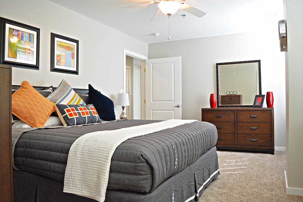 bedroom with light carpeting, bed with gray duvet, brown dresser with mirror at Residences at Forest Park, Saint Louis, MO, 63108