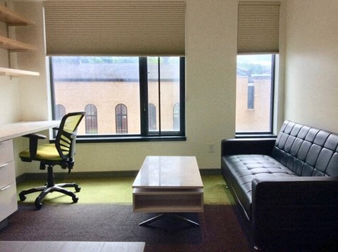 A black leather couch sits in front of a window in a room with a desk and a chair.