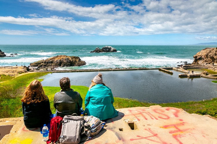 three people sitting on a ledge looking out to the ocean