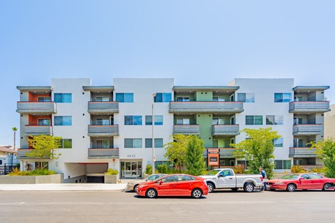 A red car is parked in front of a white building with balconies.