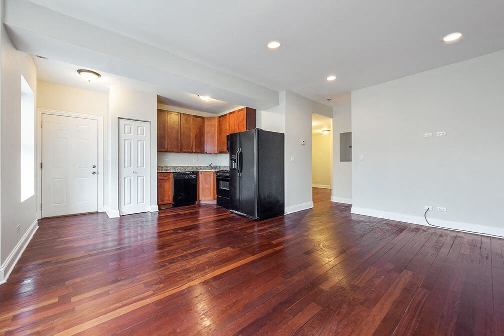 an empty living room with a kitchen with a refrigerator
