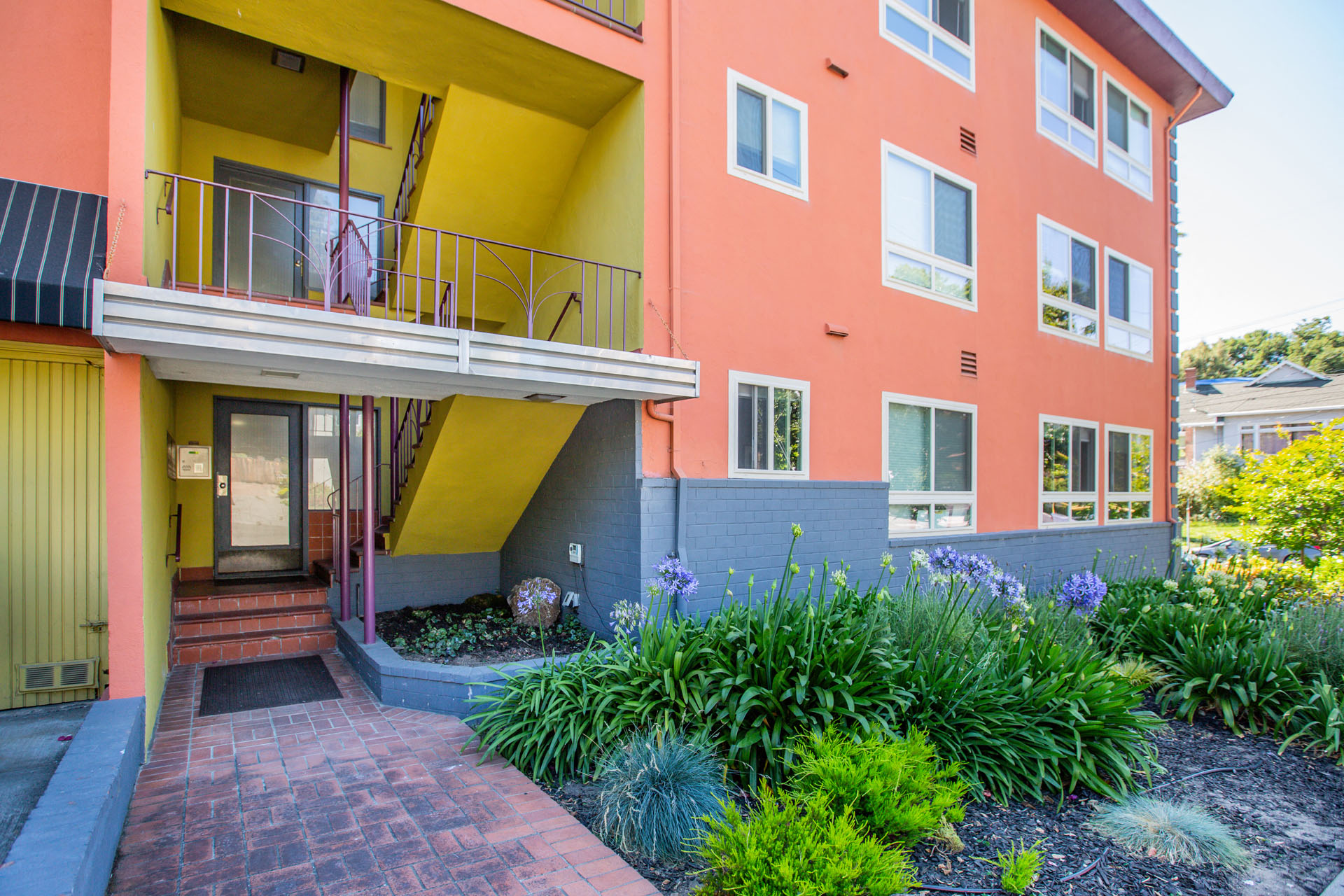 A building with a yellow and pink exterior has a balcony and a small garden in front.