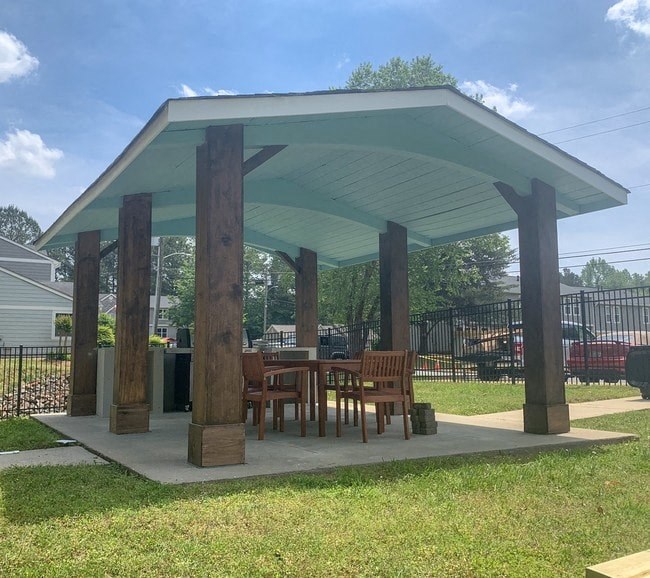 A wooden pavilion with a white roof and four pillars is surrounded by a grassy area.