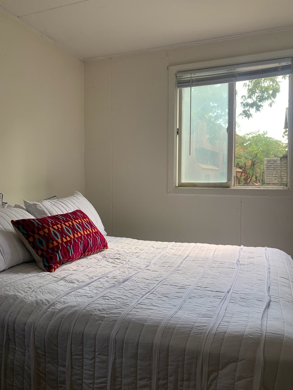 A bed with a white comforter and a red and black pillow is in front of a window.