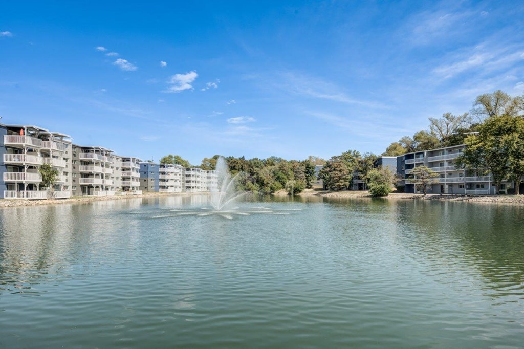 A fountain in the middle of a lake in front of apartment buildings.