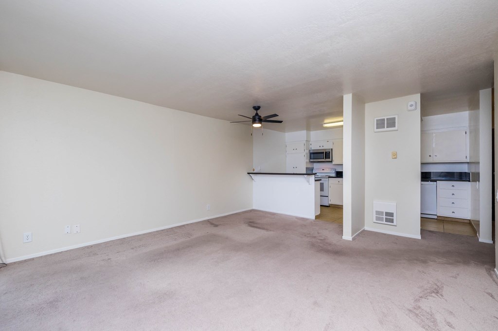 A spacious, empty room with a ceiling fan and a kitchen area visible through the open door.