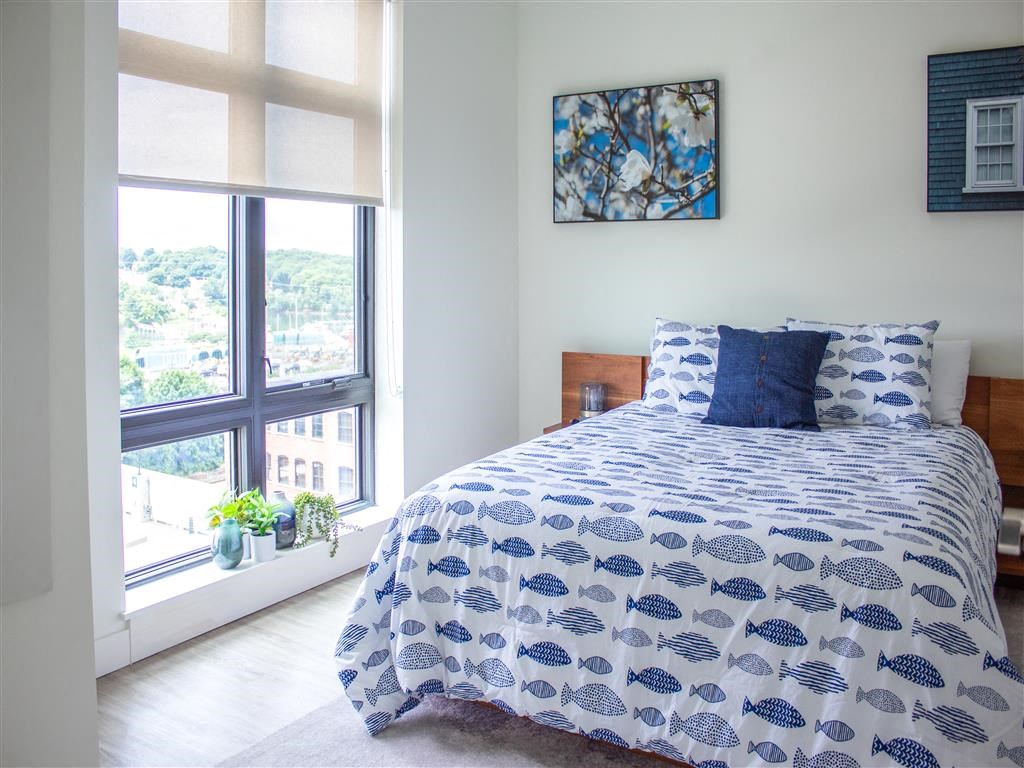1-BR Apartments in Beverly, MA - Holmes Beverly - Furnished Bedroom with Wood Flooring, a Bed, Framed Art, White Walls, LArge Window with Blinds, and Attached Nightstands