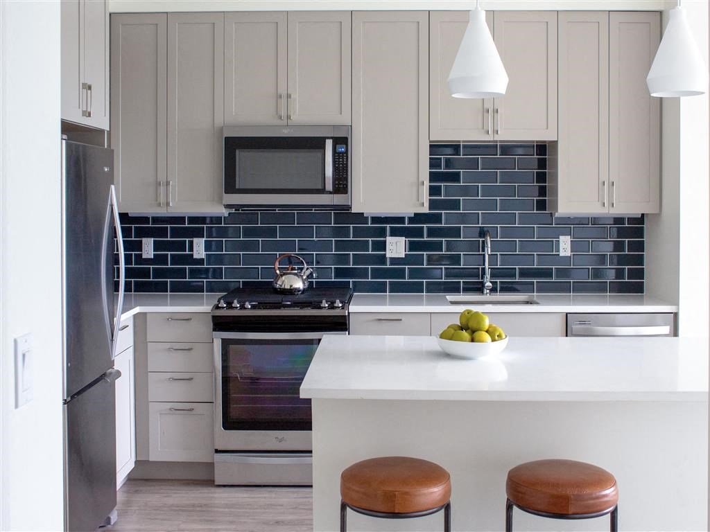 Studio Apartments in Beverly MA - Holmes Beverly - Modern Kitchen with White Countertops, Stainless Steel Appliances, Blue Tile Backsplash, Soft Grey Cabinetry, Large Cooking Island with Brown Stool Seating and Hanging Lights, and Plank Wood Flooring