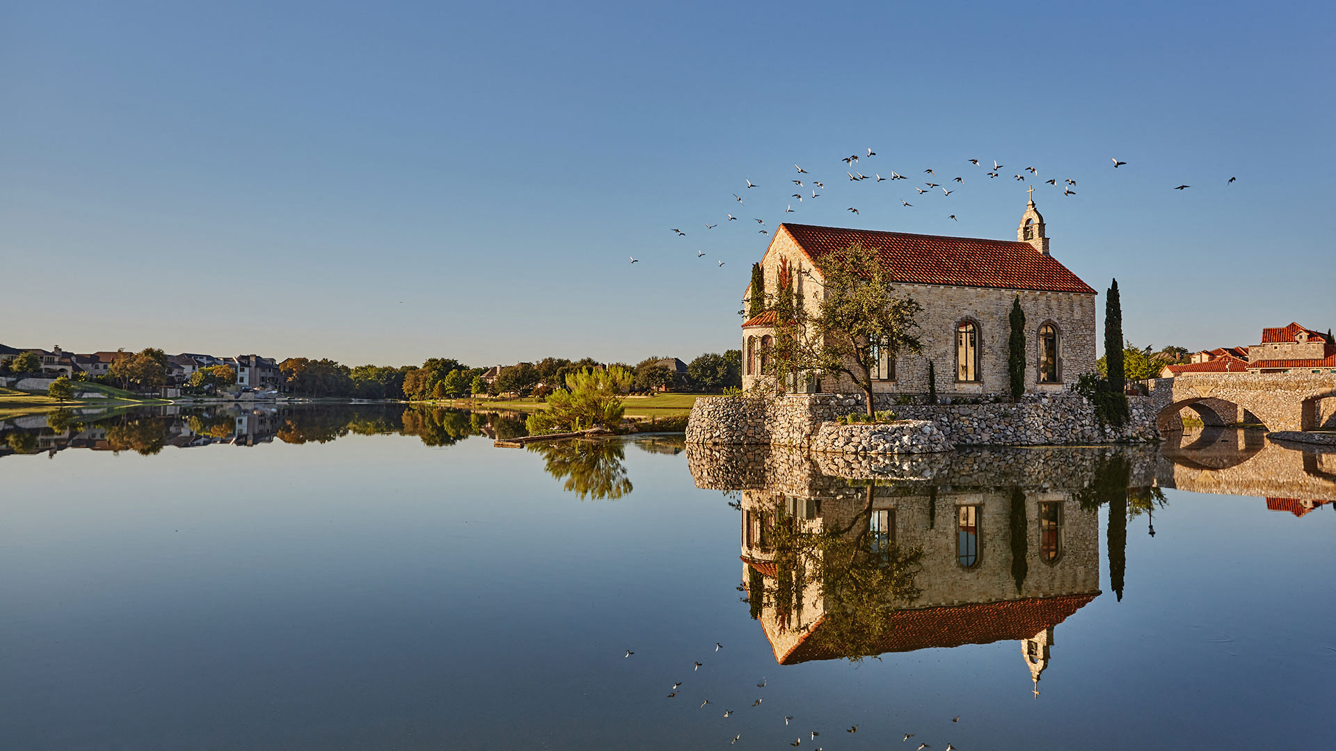 Looking across the lake at the Bella Donna Chapel at Adritica. The water is smooth, like glass and the reflection of the chapel is seen clearly. The foundation is stone as is the chapel. Trees are see