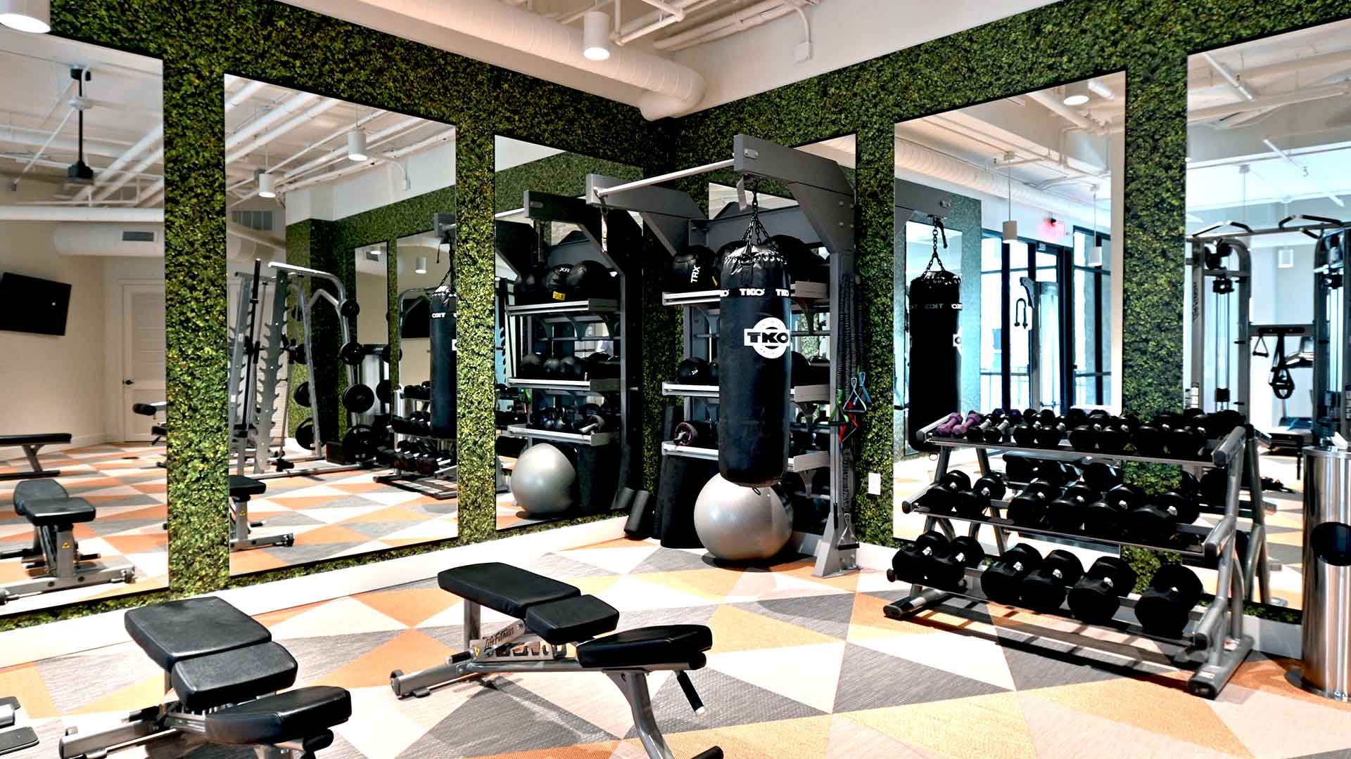 A weight bench with weights and a heavy bag to the right. The back walls are mirrored with ivy around them.
