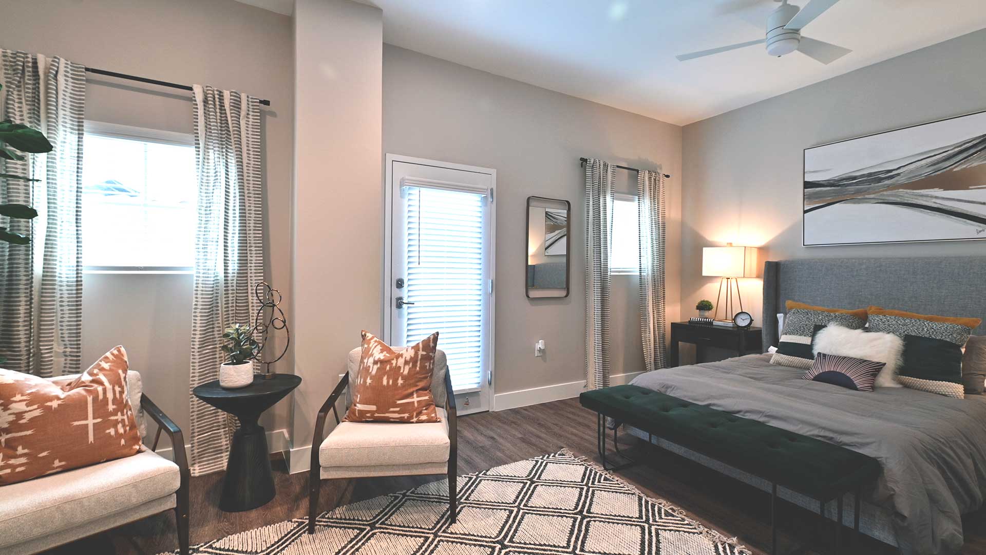 A residence bedroom at Bell Tower Flats. A large bed sits along the wall to the right, with end tables on either side. A door to the patio is to the left with a chair next to it.