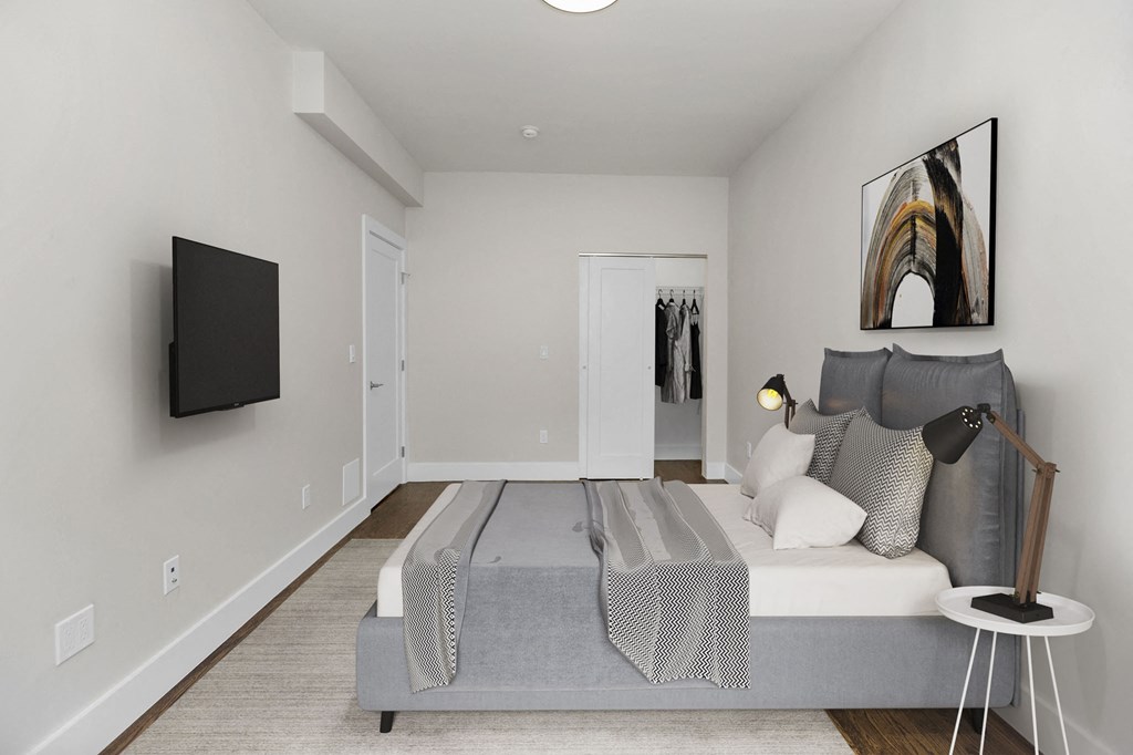 A bedroom with a bed, a grey blanket, a grey pillow, a black lamp, a black television, a white wall and a white door.