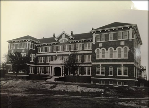 A black and white photo of a large, two-story building with a steeply pitched roof and a cross on the top.