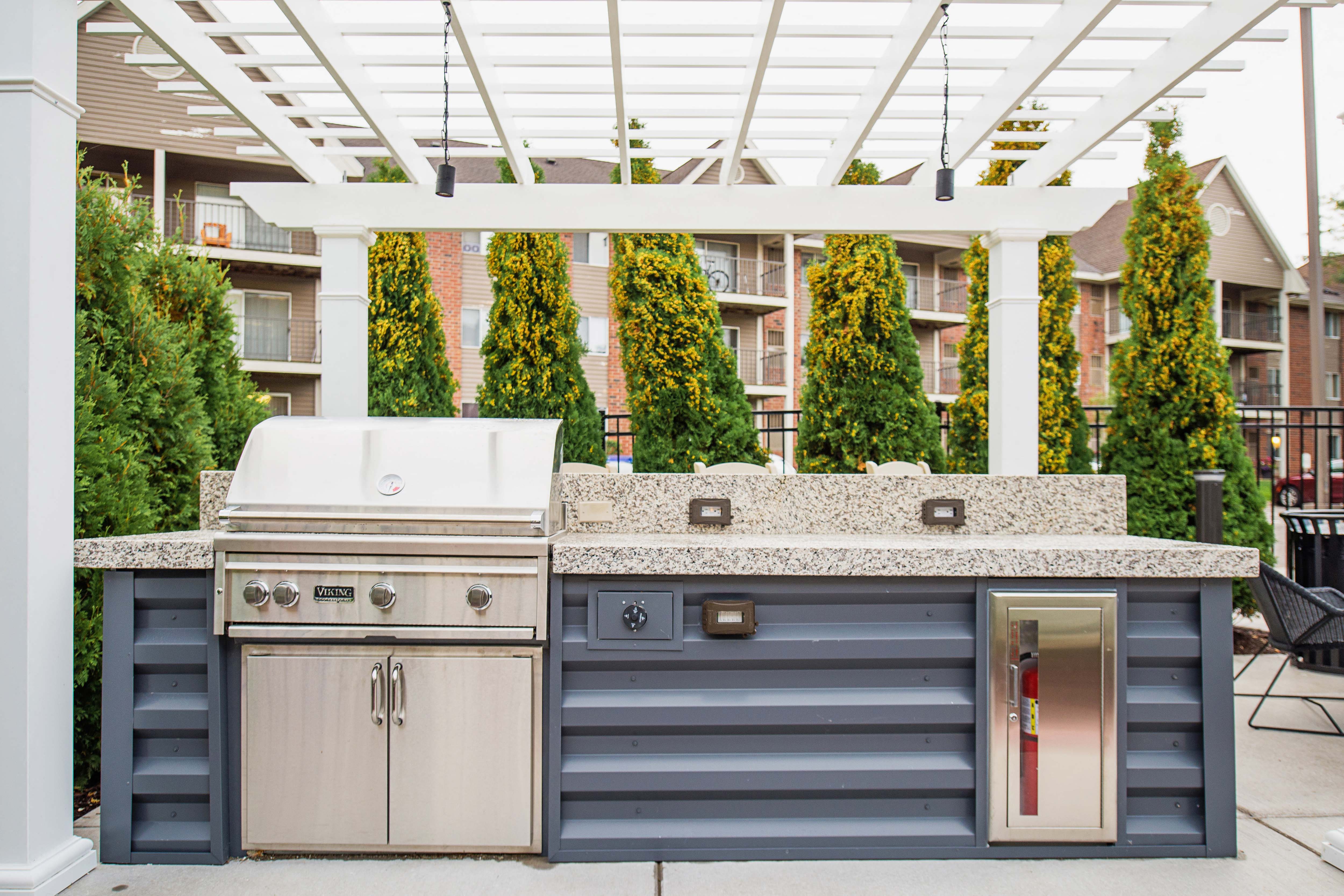 A communal outdoor patio adorned with ample seating arrangements, a cozy gas fireplace, and a fully-equipped grill ready for summer cookouts. The atmosphere radiates warmth and relaxation, offering residents a perfect setting to unwind and socialize amidst the comfort of their living space.