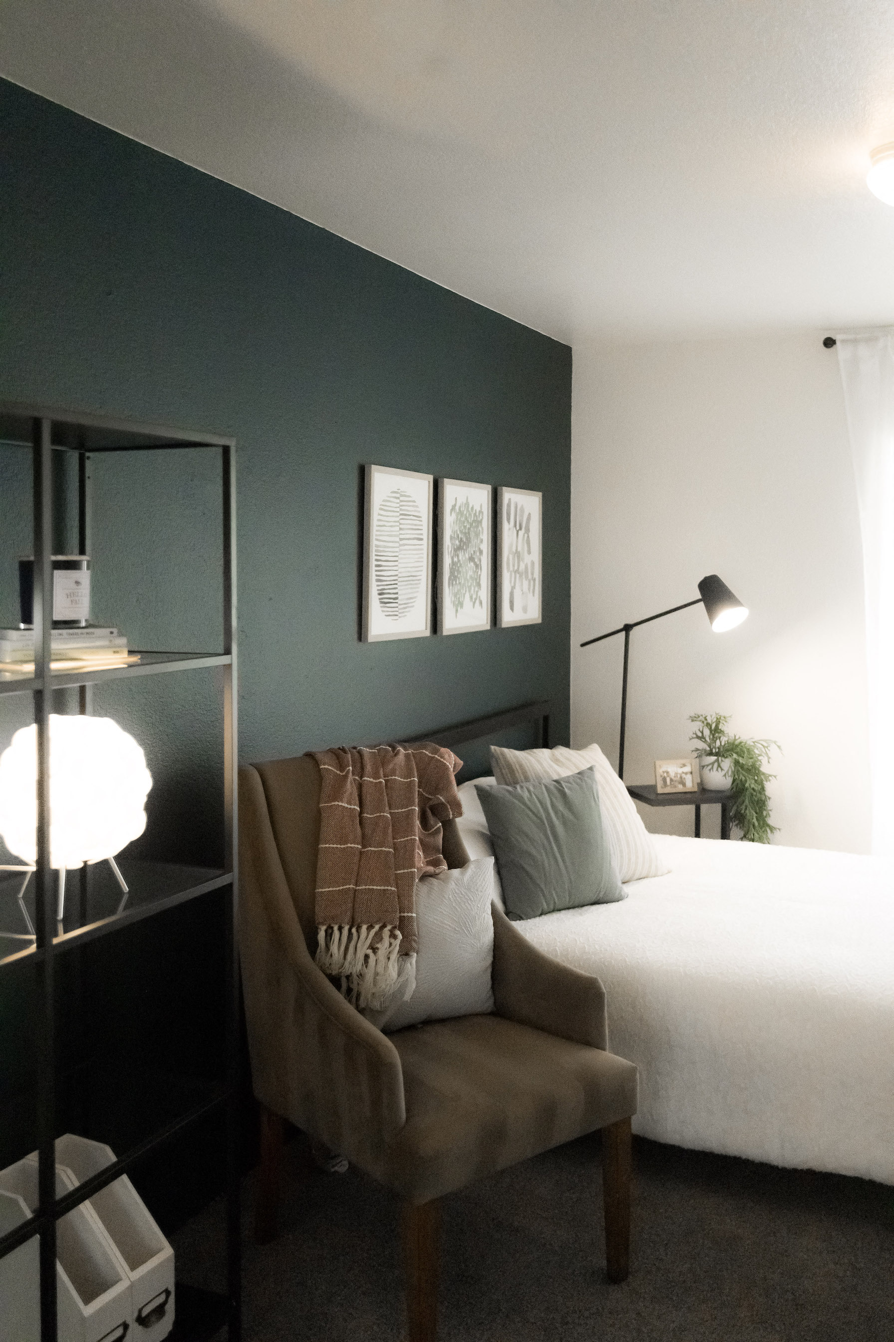 Image of a room with a dark blue, green wall with a bed, chair, and shelf in view