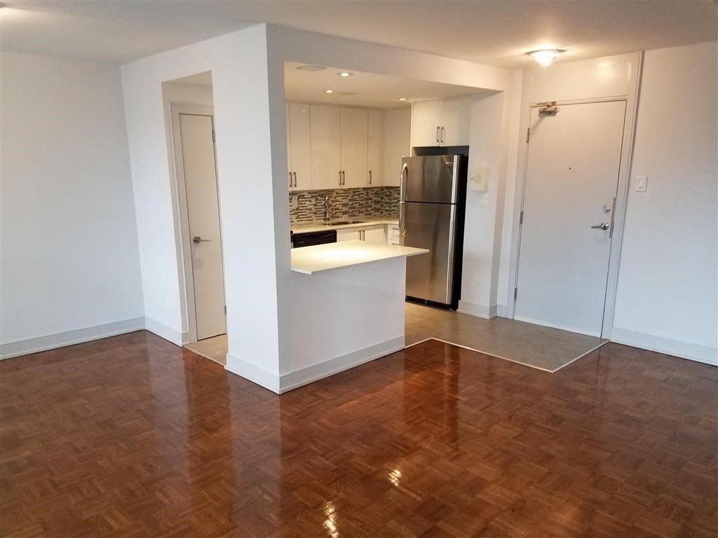 an empty living room with a kitchen with a refrigerator