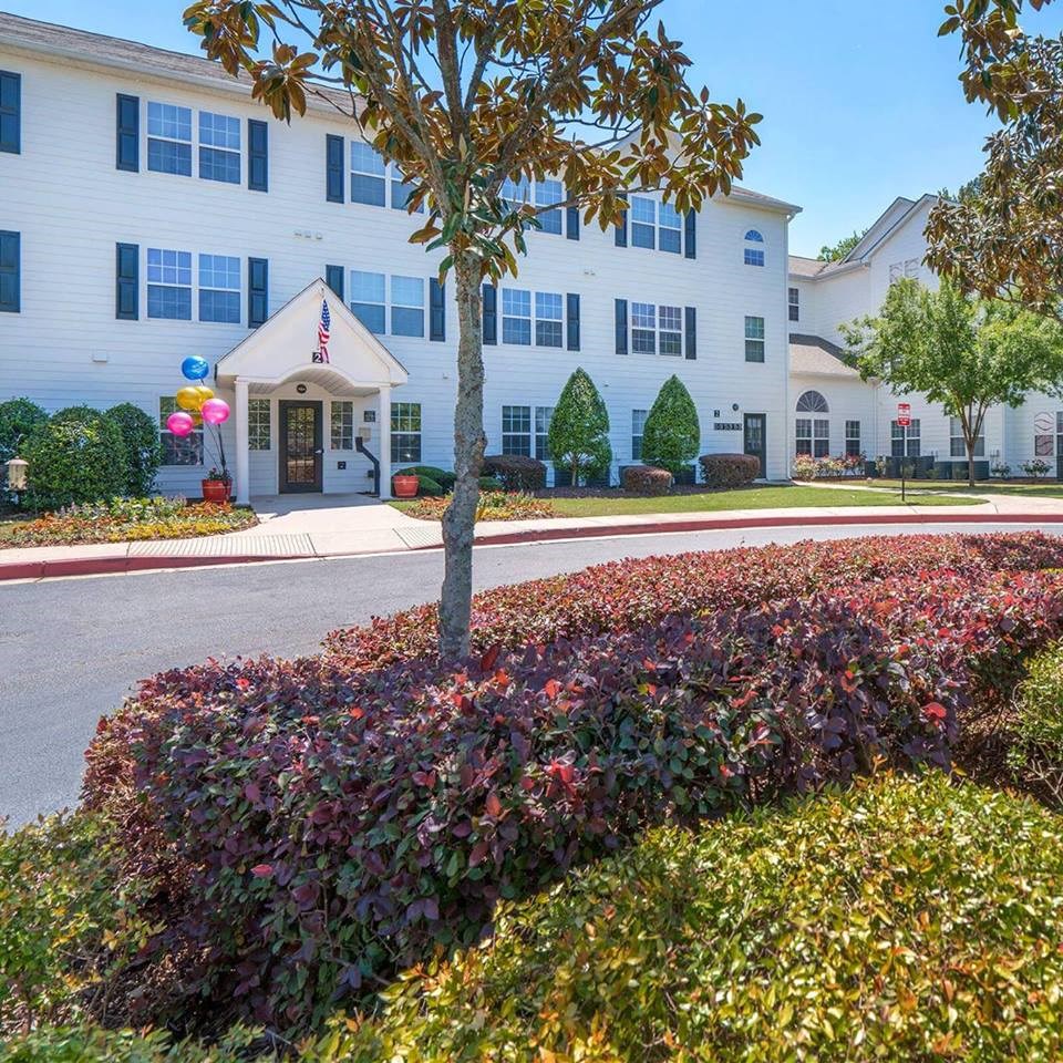 A well-maintained three-story white residential building with black shutters, surrounded by lush landscaping. A small tree and neatly trimmed bushes are in the foreground. The entrance has a white gabled awning with an American flag and colorful balloons tied near the doorway. A clear blue sky is visible above