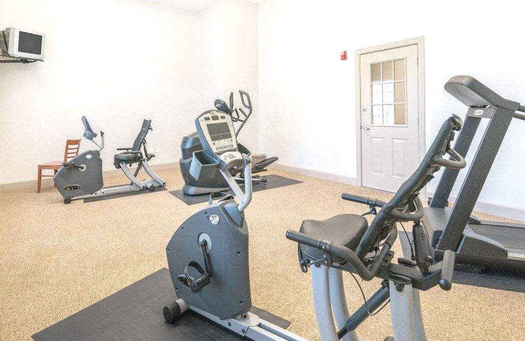 Apartment gym with a treadmill, two stationary bikes, and an elliptical on a carpeted floor. A wall-mounted TV is in the corner, and a door with a glass panel is on the right side. The room has neutral-colored walls and bright lighting.