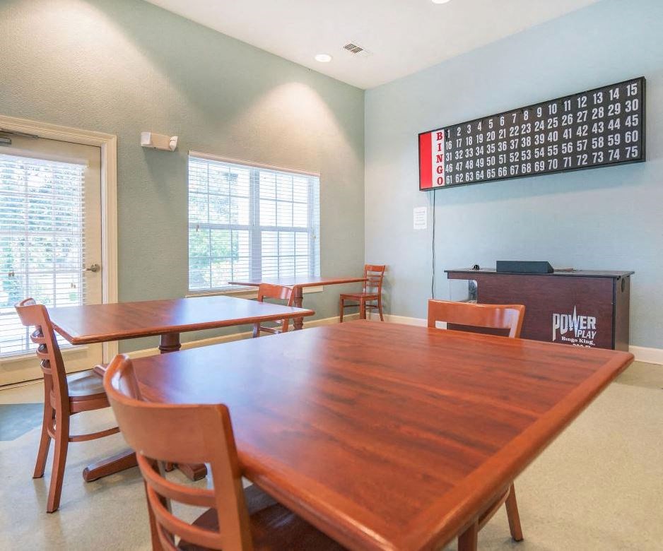 A community game room with wooden tables and chairs arranged for seating. A large bingo board is mounted on the wall above a desk labeled 'Power Play Bingo King.' Natural light enters through windows and a glass door with blinds