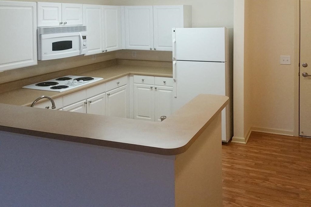 A kitchen with white cabinets, beige countertops, and wood flooring. It features a white refrigerator, microwave, and electric stovetop. A raised breakfast bar separates the kitchen from the living space