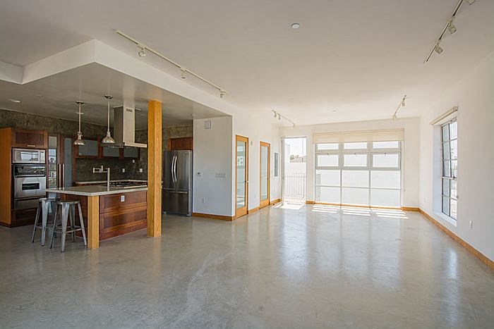 A large, empty room with a kitchen area to the left and a doorway leading to another room to the right.