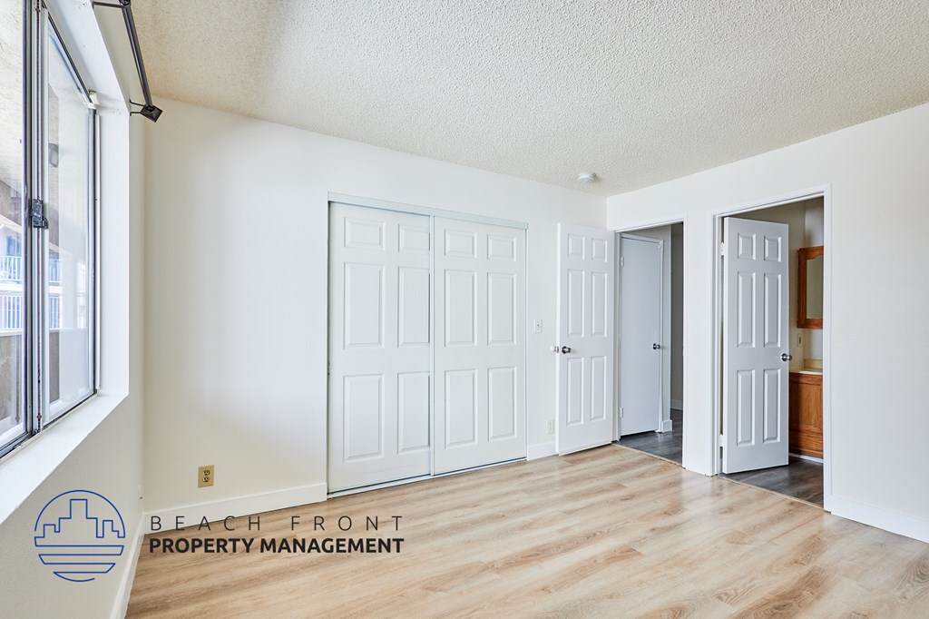 A room with white walls and wooden floors, featuring a white door and a window with a view of buildings outside.