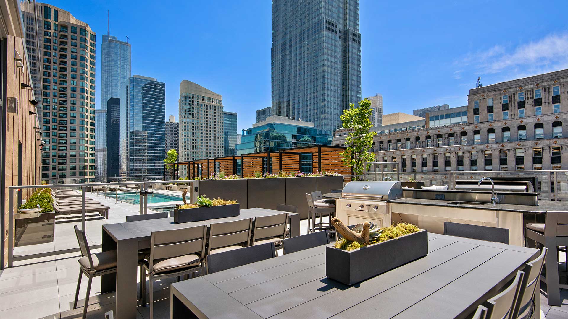 Looking across an outdoor dining table at a grilling station behind it. The pool deck is off to the left. City buildings rise in the background on a clear day.