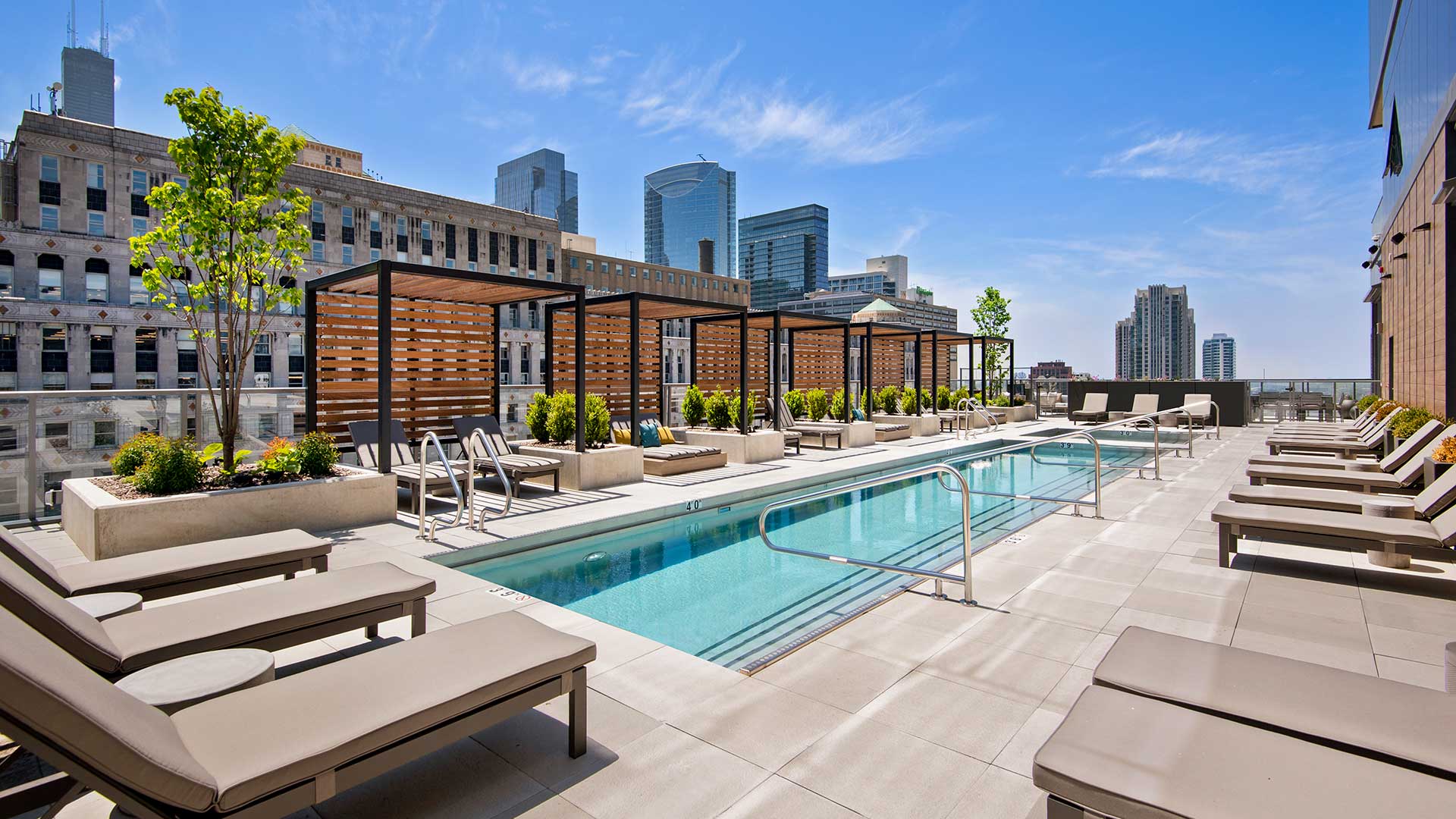 Looking across the outdoor pool at Hubbard221. Lounge chairs go off to the left and right. Pergolas are across the pool with more lounge chairs beneath them. City buildings are seen rising in the back