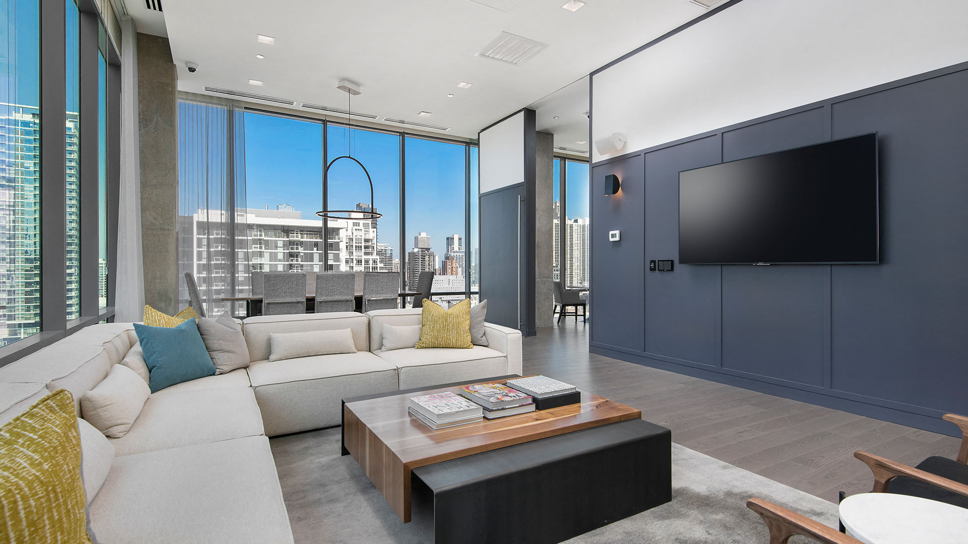 Looking from the perspective of a chair in the sitting area of the part room, a sectional goes off to the left, a dining table set behind it. To the right is another chair and a television mounted on