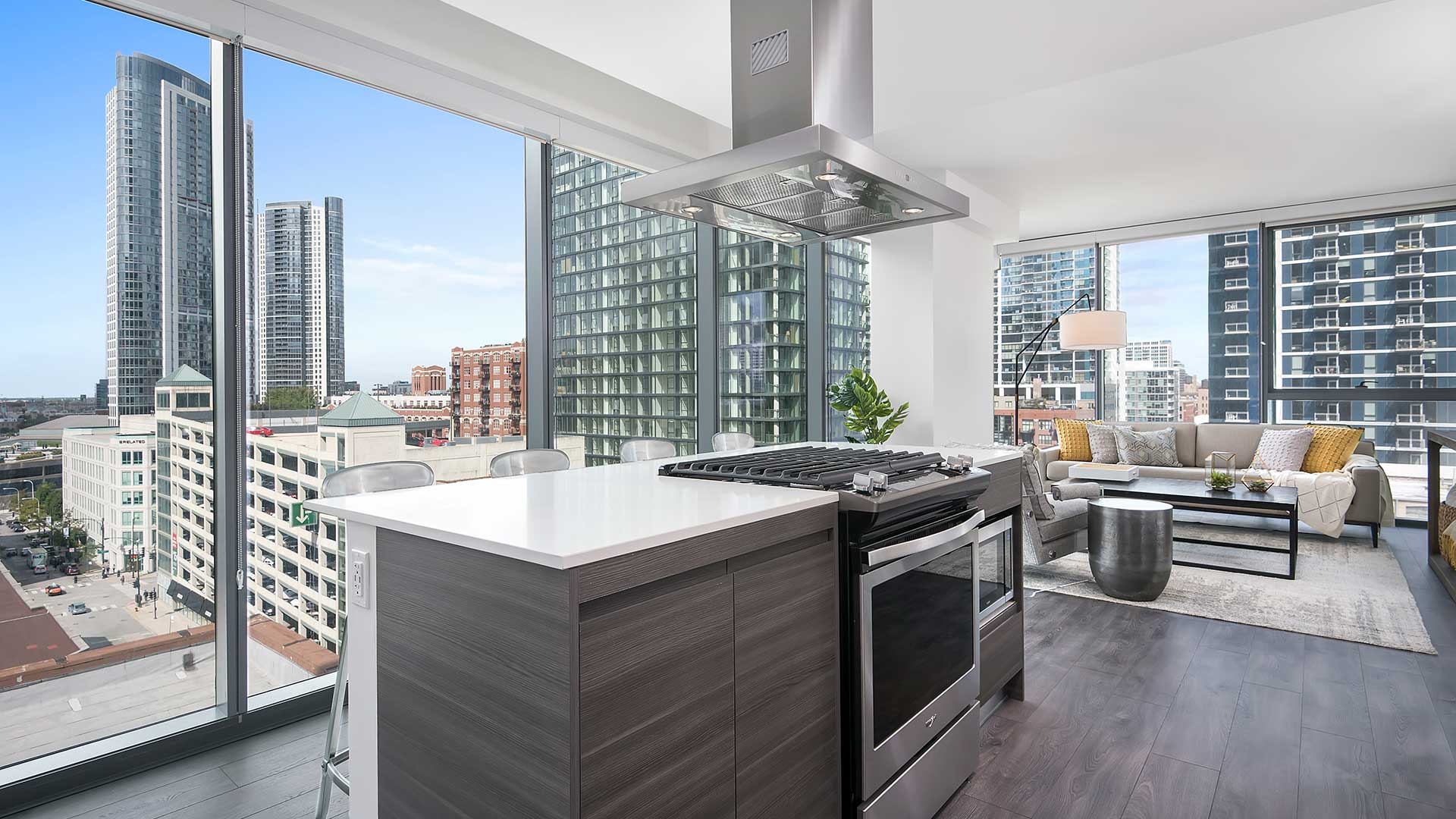 Looking across a kitchen island in a Hubbard221 residence. There is a stainless steel stove with range in the island, a stainless steel exhaust hood above. Beyond the kitchen to the right is a living