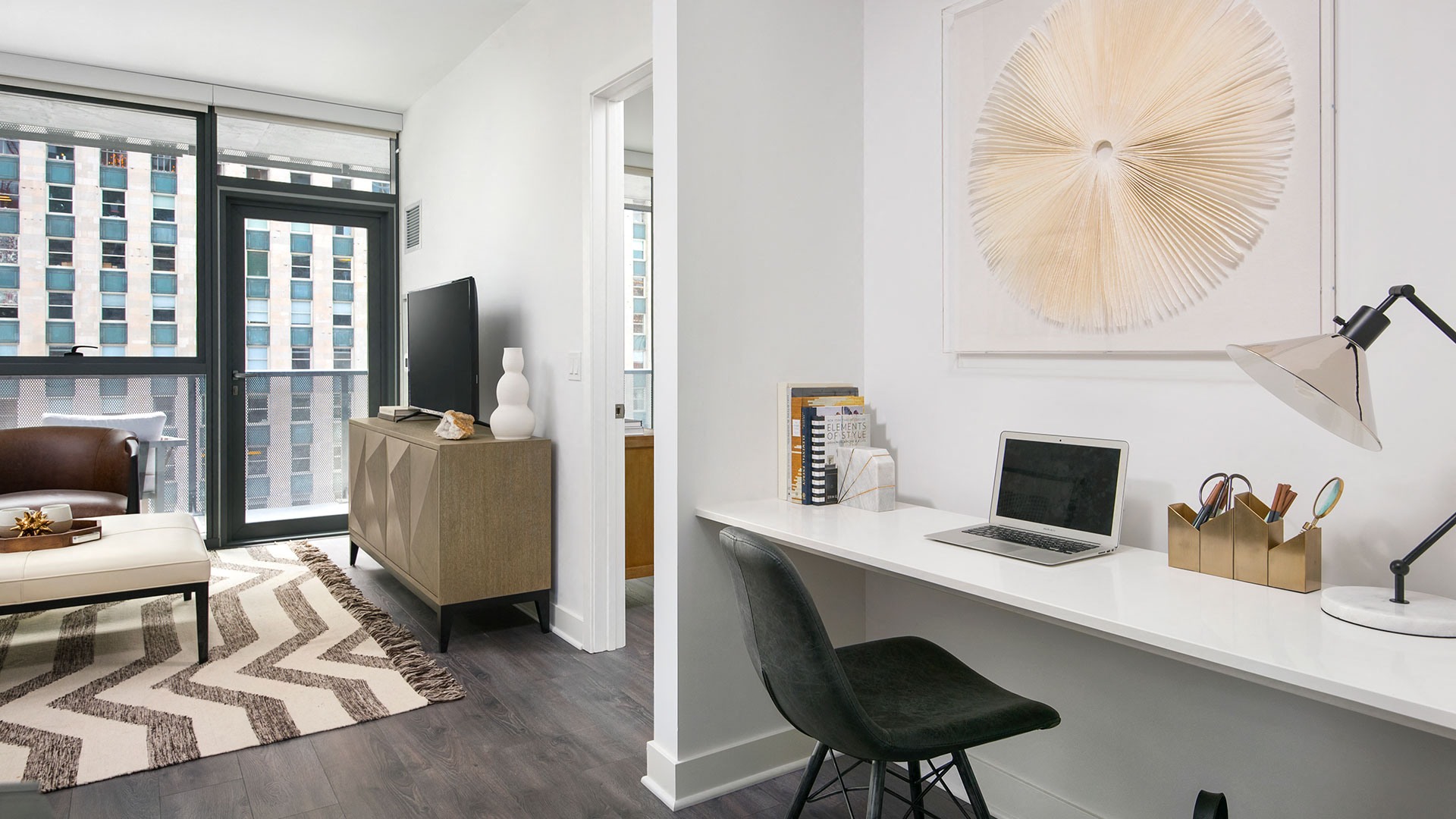 A built-in desk is on the wall on the right in this residence. Just past is a doorway to the bedroom. On the left is a living area with couch and television.