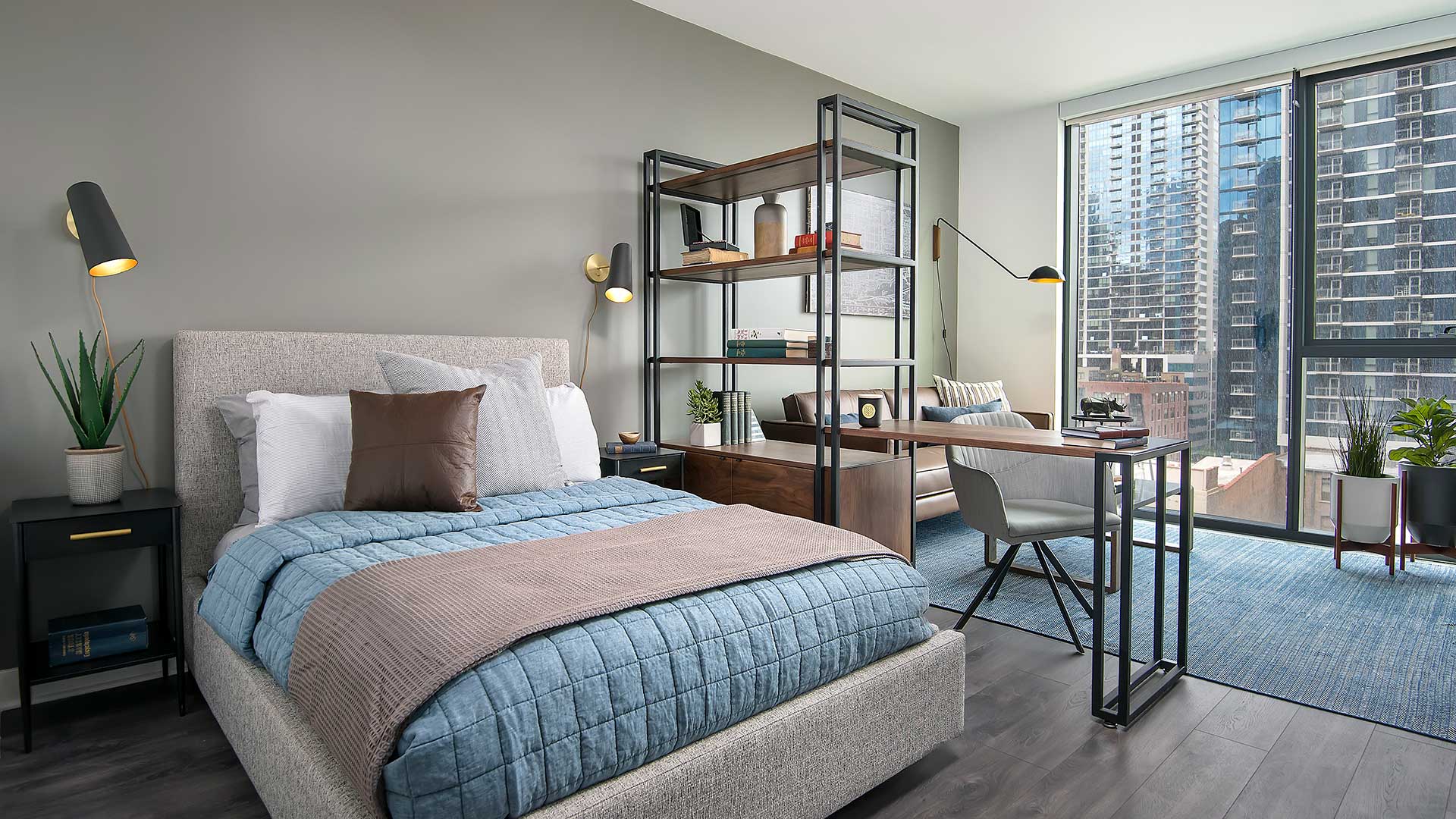 A studio residence with the bed on the left, an end table on each side. A framed bookcase separates the room in the middle with a desk and living space beyond it. Floor-to-ceiling windows are at the b