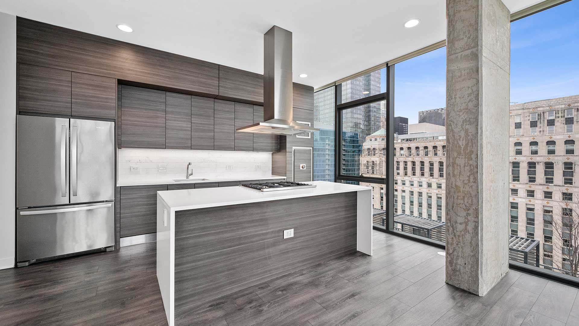 Looking into a kitchen in a residence at Hubbard221. The kitchen is on the left, the back lined with cabinets, a counter and fridge. A kitchen island is in front with a range and range hood overhead.