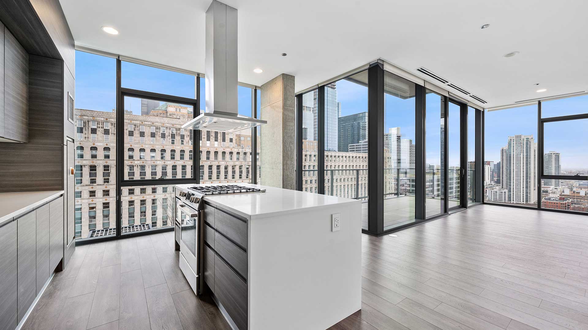 An empty unit with floor-to-ceiling windows and a balcony along the far walls. In the foreground is a kitchen island with an oven and range top built-in, a range hood hangs overhead.