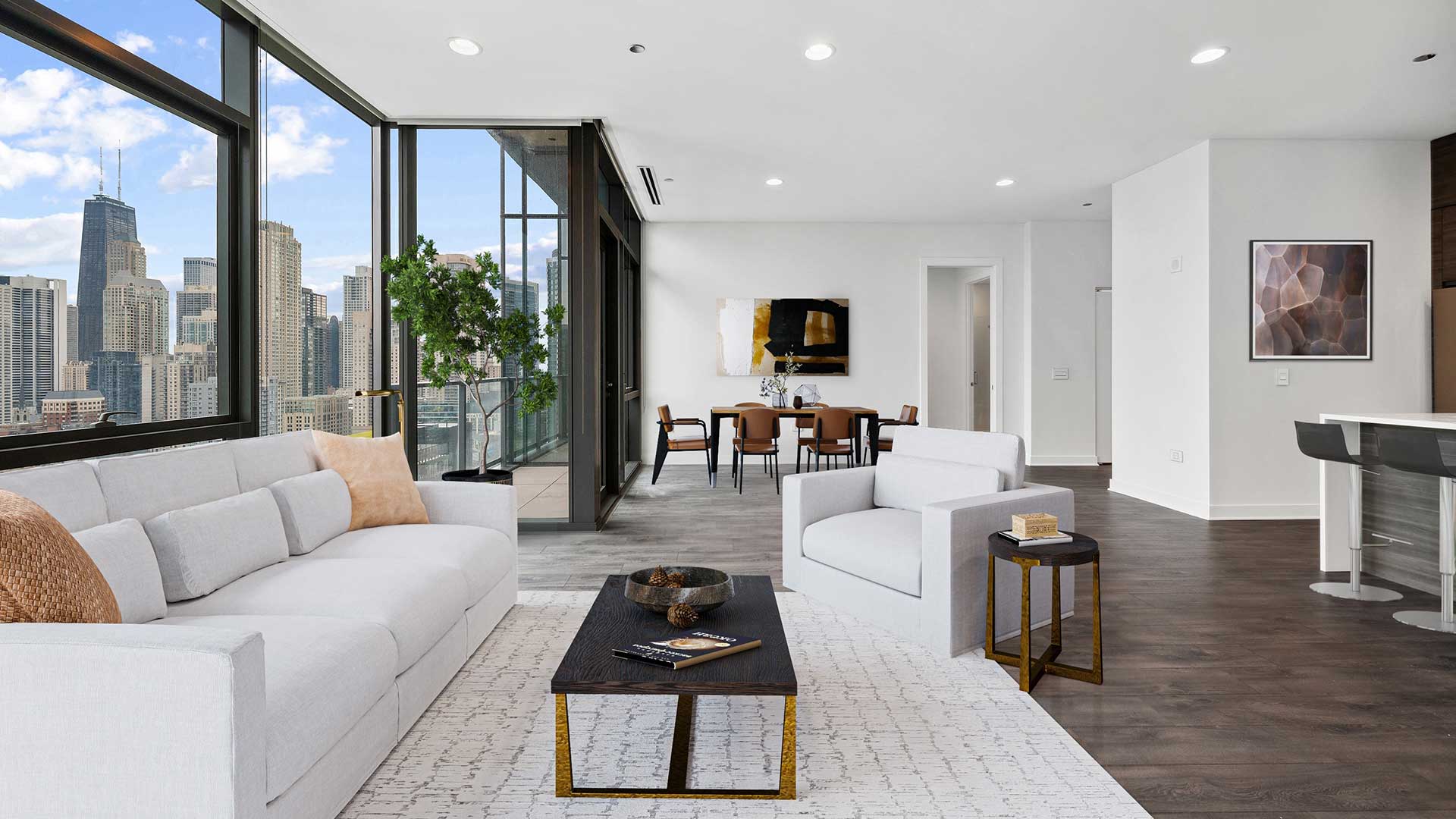A large living room in a Hubbard221 residence. There is a living room set in the foreground with a couch, chair and coffee table. Beyond is a dining set. The left are floor-to-ceiling windows that sho