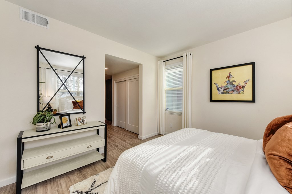 Master bedroom with white bed and copper colored throw pillows, Black and white dresser with mirror positioned above it at Morgan Ranch Apartments, Morgan Hill