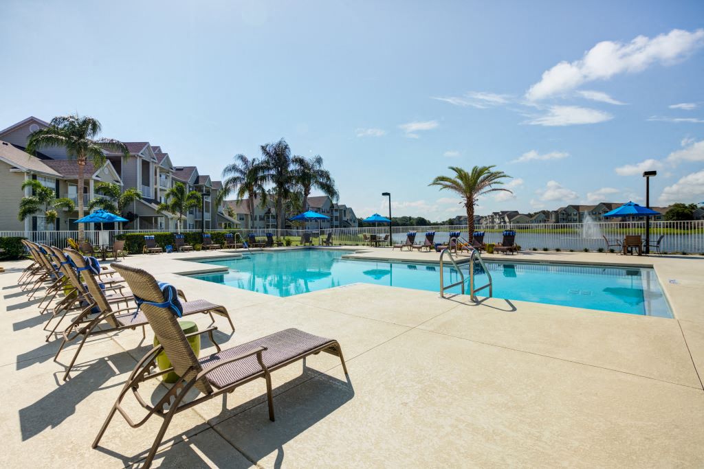 Apartments for Rent in Lakeland, FL - Village at Lake Highland - Communal Pool with Blue Waters, Lounge Chairs, White Fence, with View of Pond with Fountain, and Surrounding Palm Trees and Greenery