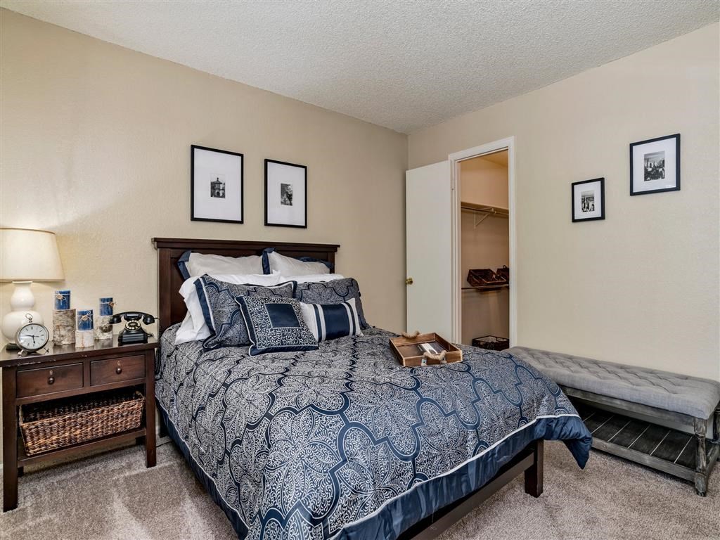 Bedroom with a dark wooden bed frame, navy and white patterned bedding, and decorative pillows. A nightstand with a lamp, clock, and framed photos sits beside the bed. A gray upholstered bench is against the wall, and an open door leads to a walk-in closet.