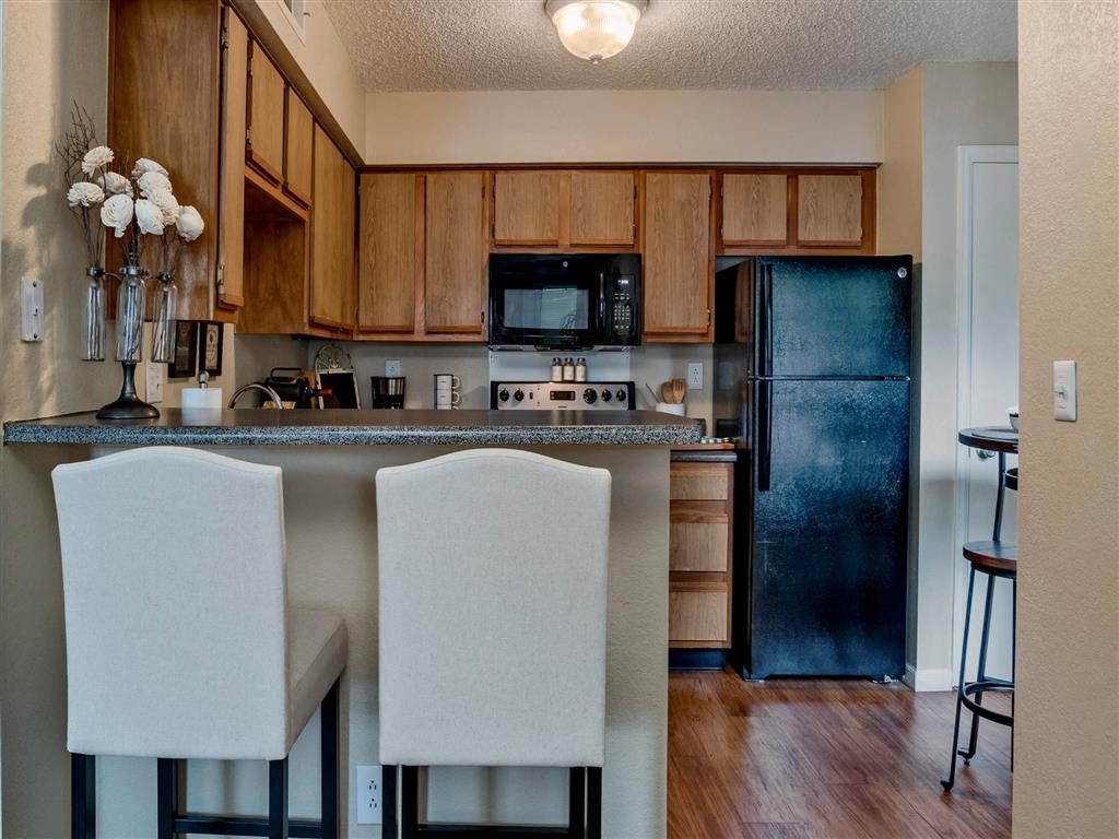 Modern kitchen with wooden cabinets, black appliances, and a granite countertop. Two upholstered barstools sit at the breakfast bar, and a small dining table with chairs is visible in the background. Decorative flowers and kitchen essentials add a cozy touch