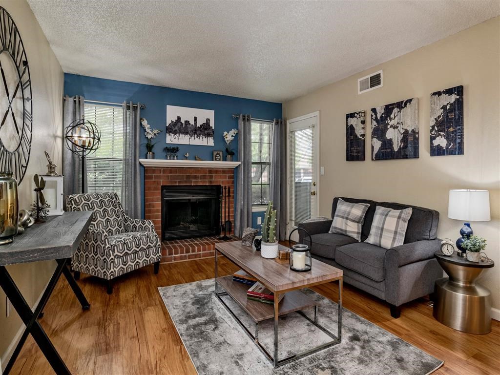 Cozy living room with a brick fireplace, blue accent wall, and modern furnishings. Features a gray loveseat with plaid pillows, a geometric-patterned armchair, a wooden coffee table with decor, and large windows with gray curtains