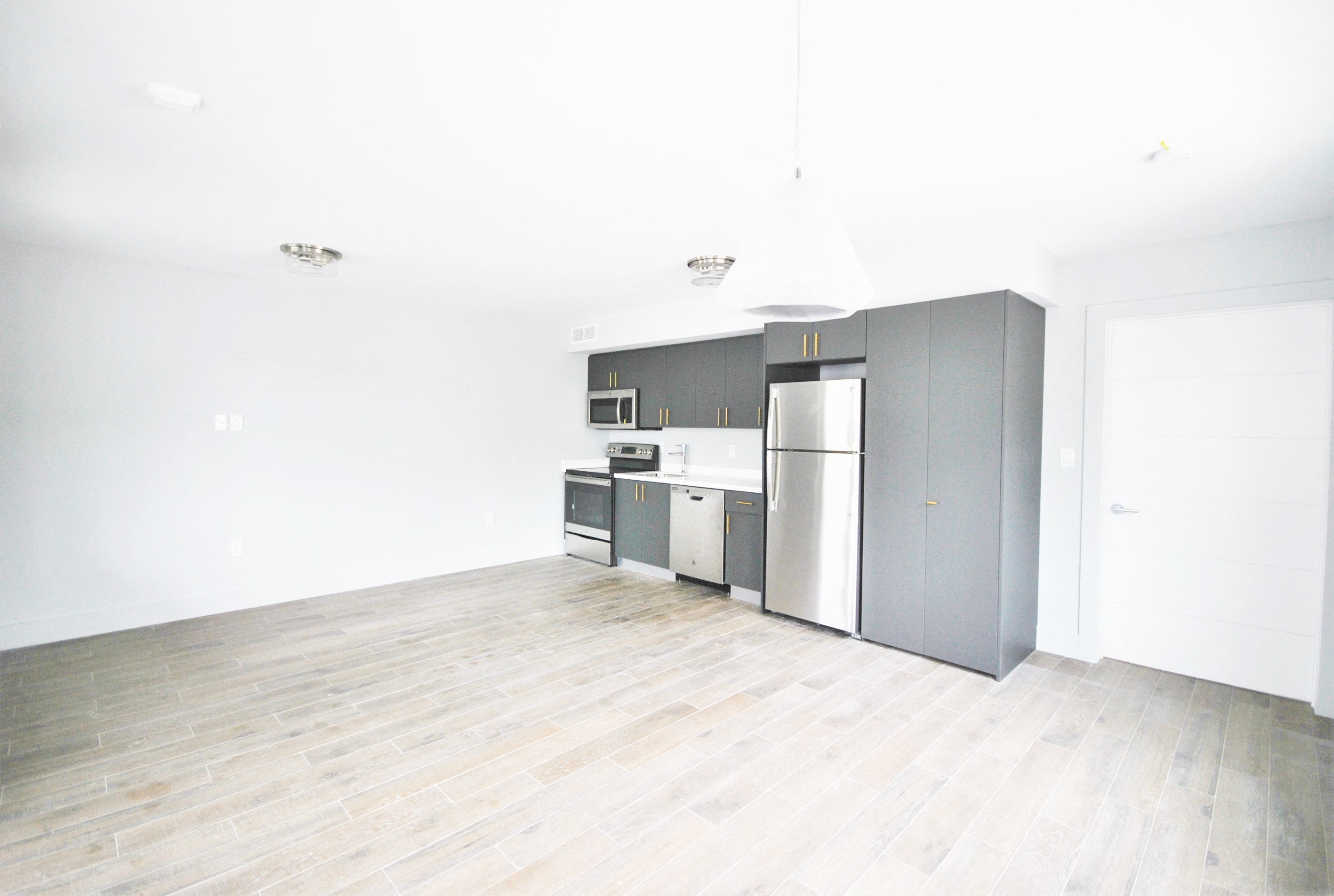 an empty living room with a kitchen with a refrigerator and