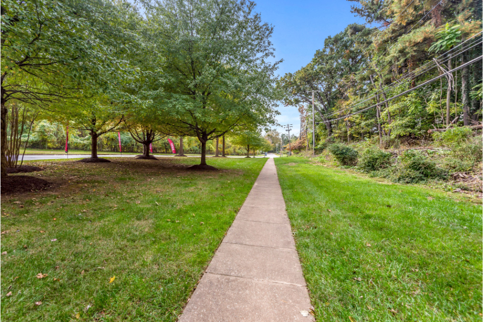 A stretch of sidewalk with green grass on both sides. There is a fence and some shrubbery to the right, and tall trees with green leaves to the left