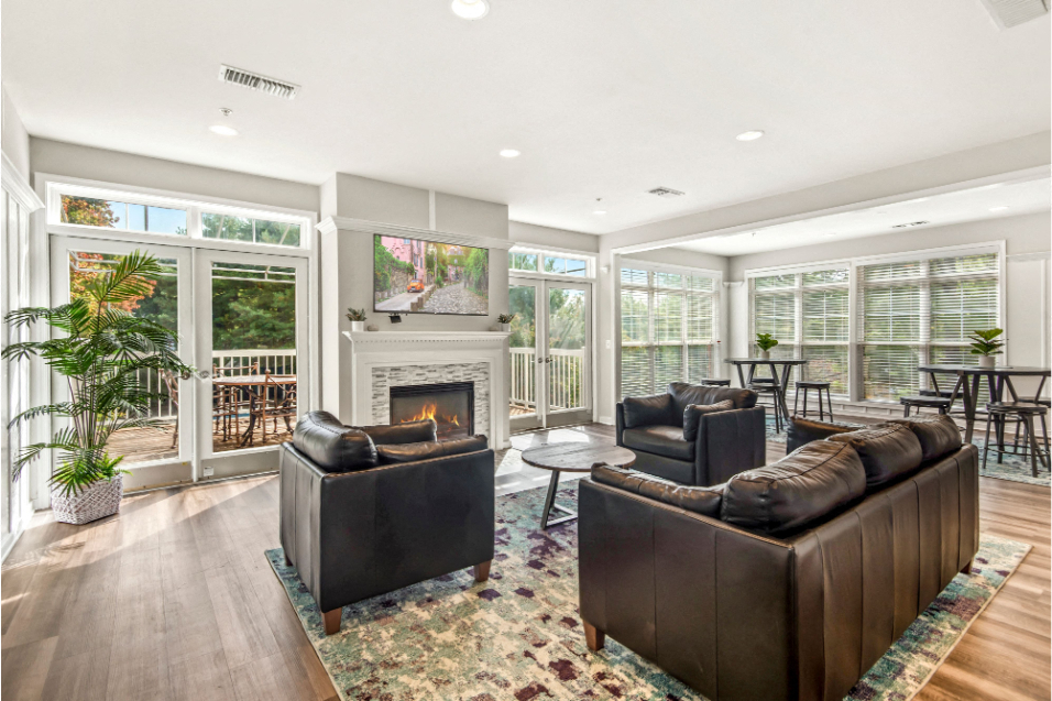 A room with white walls, floor-to-ceiling windows, and medium-brown wood floors. There is a rug in the middle of the room, with leather couches and a small coffee table across from a lit fireplace