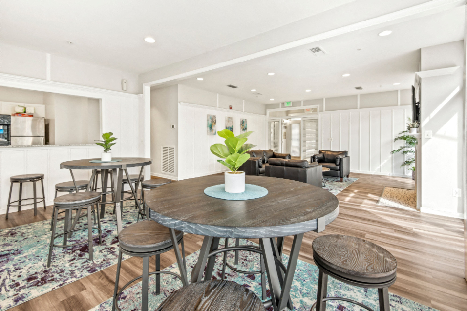 A room with white walls, floor-to-ceiling windows, and medium-brown wood floors. There is brown leather furniture by a fireplace on one end of the room, and tall pub tables with stools at the other