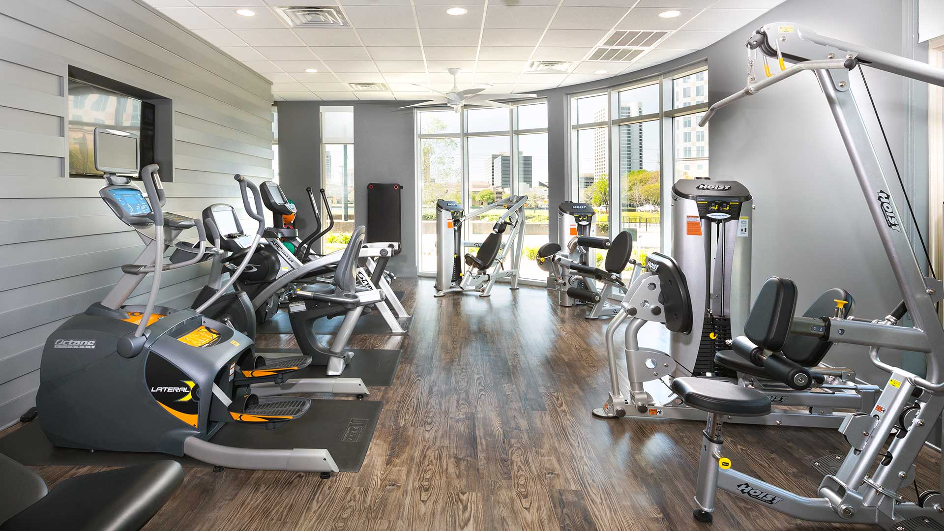 Looking down a row of various fitness machines with windows looking outside at the far wall.