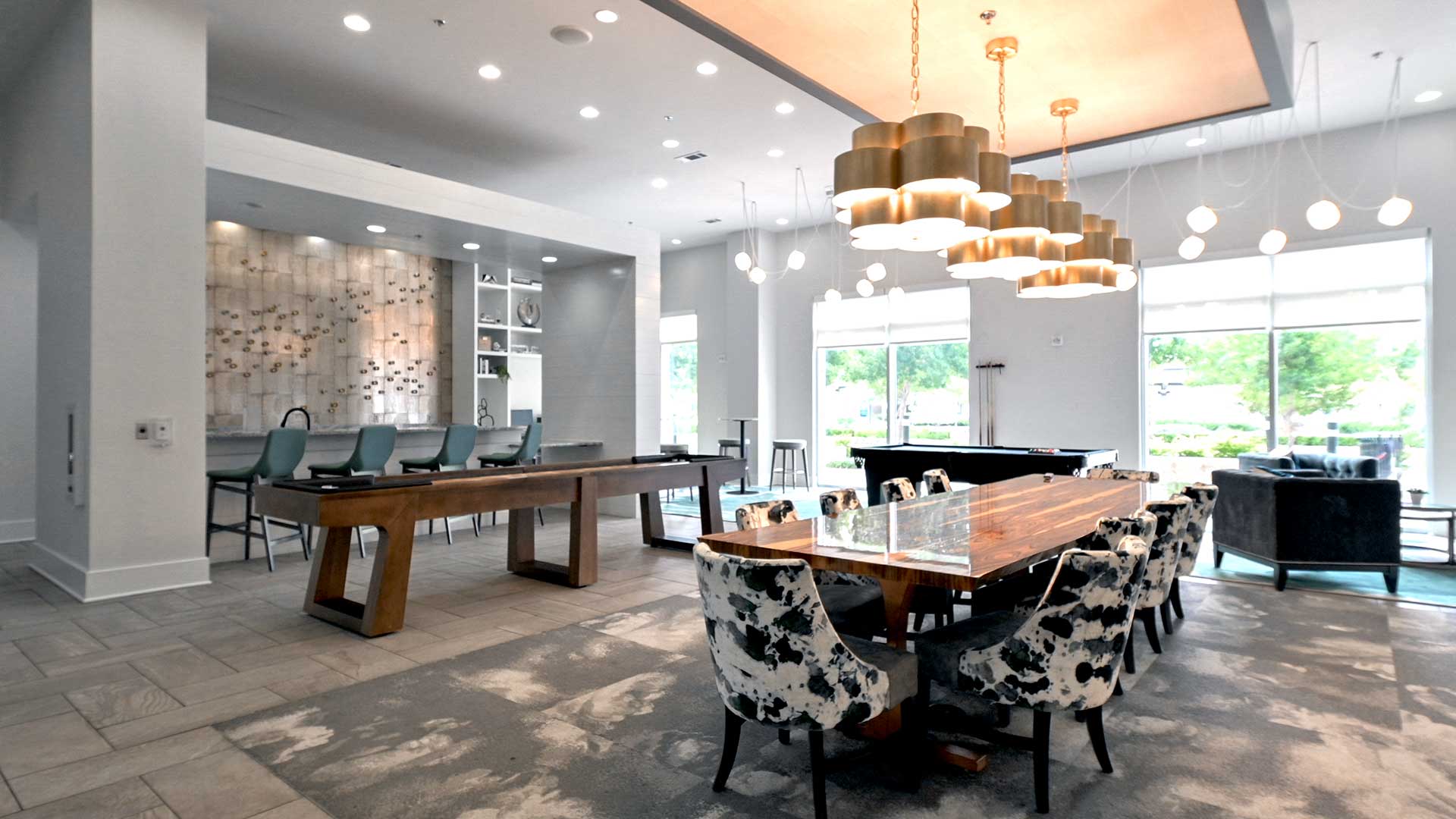 A large dining table with pattered chairs sits below three, large chandeliers. Behind is pool table and shuffleboard table. The far left wall is a bar and the back wall is a set windows to the pool de